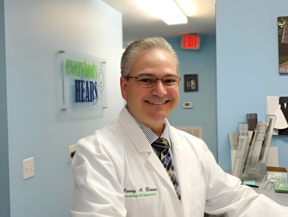 A man in a white lab coat and glasses smiling in an office with a sign on the wall and a stack of folders.