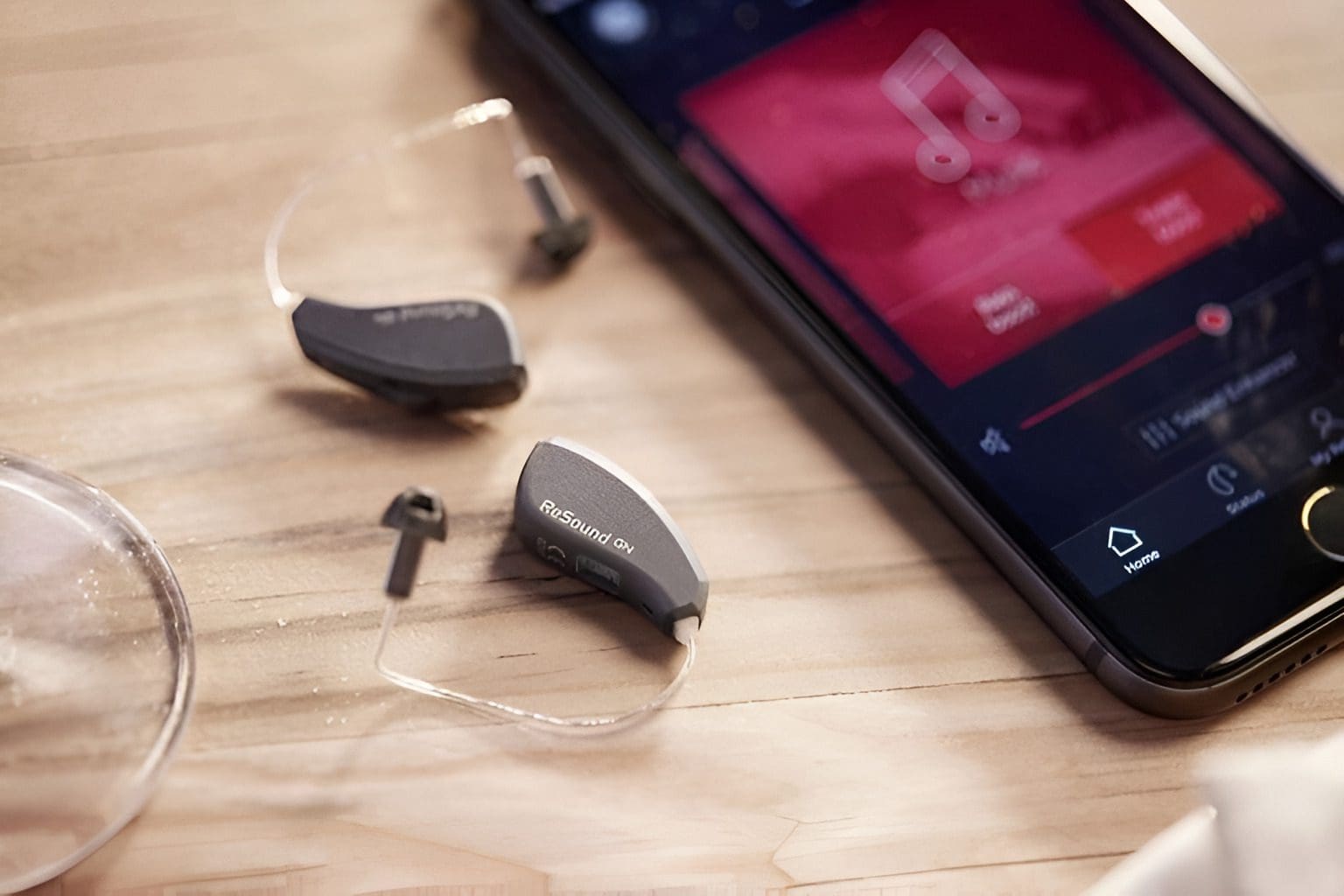 Hearing aids placed on a wooden surface next to a smartphone displaying a music app.